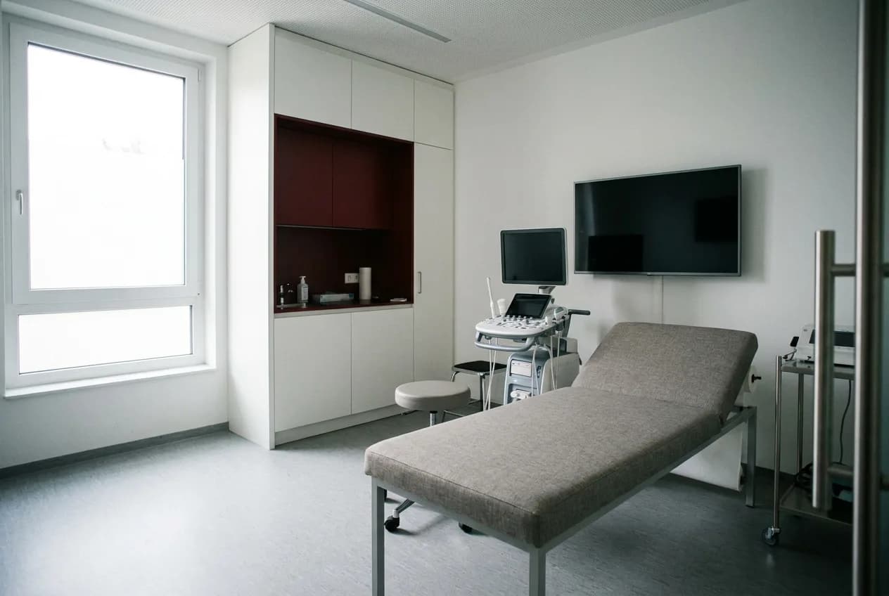 Heller Behandlungsraum mit Untersuchungsliege, Ultraschallgerät und Wandmonitor, Tageslicht durch ein großes Fenster.